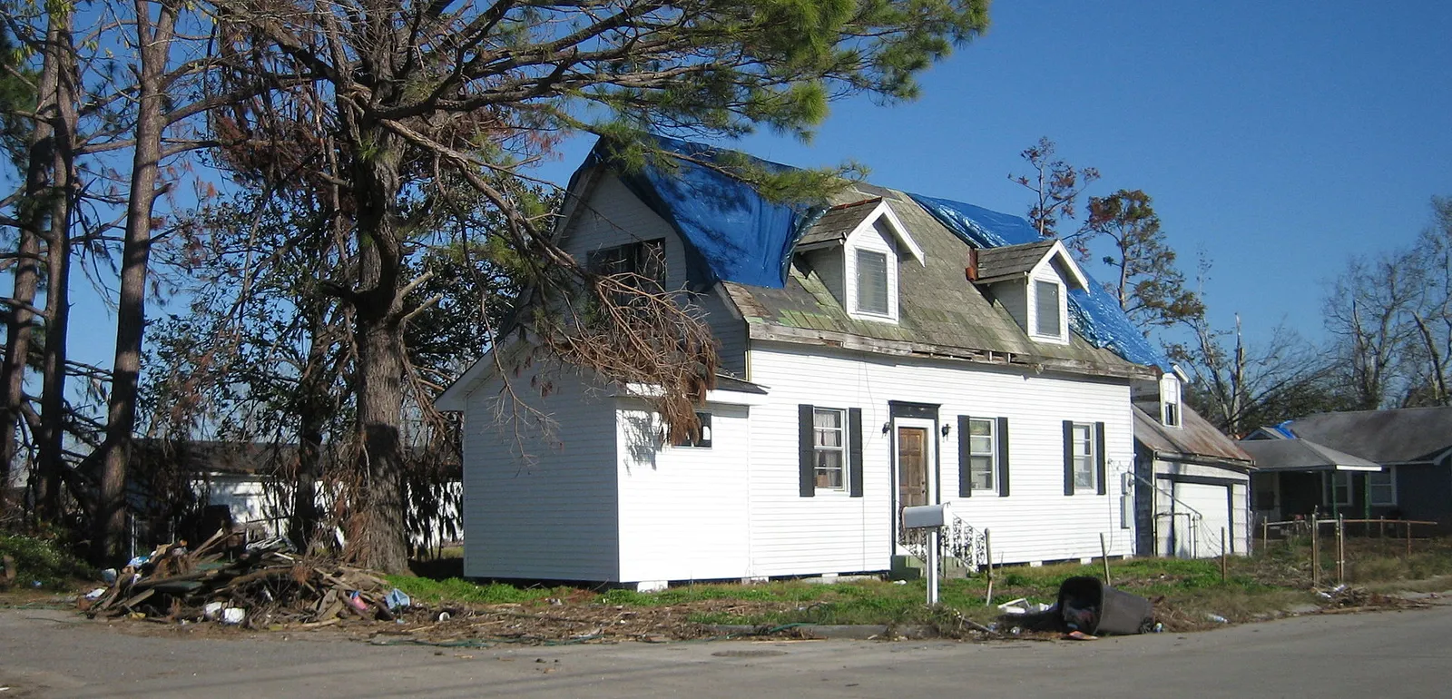 A house in New Orleans East with a blue tarpaulin over its storm-damaged roof and debris piled at the kerb