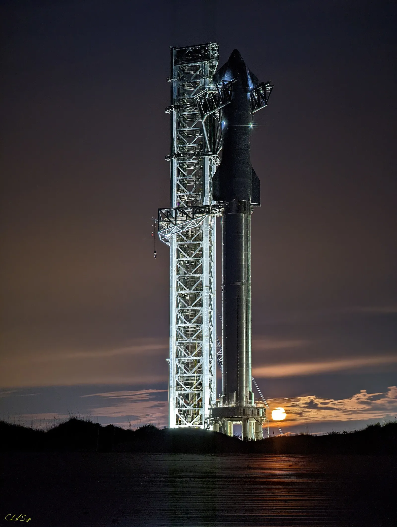 A SpaceX Starship rocket on the pad with the moon setting behind it